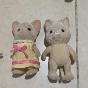 Puede incluir: Dos peque&ntilde;as figuras de animales flocados. Una es un rat&oacute;n que lleva un vestido amarillo con un lazo y ribete rosa. La otra es un gato. Ambos tienen ojos negros y orejas forradas de rosa. Las figuras son de color beige y marr&oacute;n.