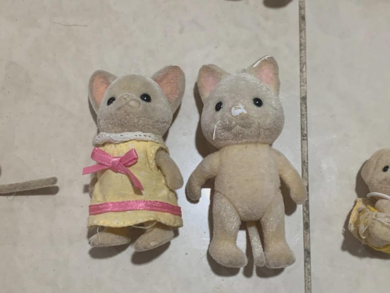 Puede incluir: Dos peque&ntilde;as figuras de animales flocados. Una es un rat&oacute;n que lleva un vestido amarillo con un lazo y ribete rosa. La otra es un gato. Ambos tienen ojos negros y orejas forradas de rosa. Las figuras son de color beige y marr&oacute;n.