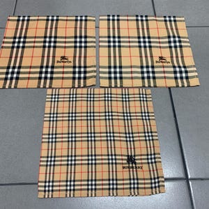 May include: Three tan, red, black, and white plaid handkerchiefs. Each handkerchief features the Burberry logo in black. The handkerchiefs are arranged on a gray tiled surface.