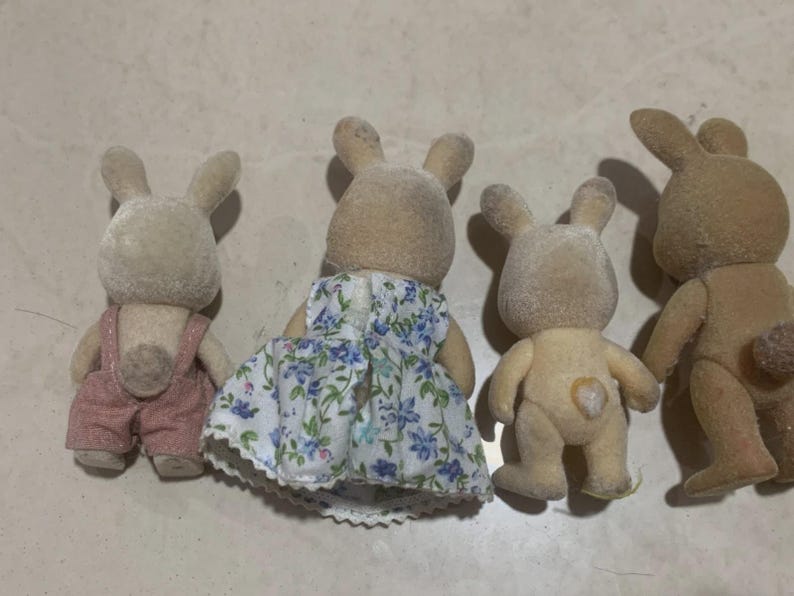 Puede incluir: Cuatro figuras de conejos de juguete. Un conejo lleva un peto rosa, otro un vestido floral. Los otros dos conejos son de color beige natural. Los juguetes tienen orejas largas y est&aacute;n de pie.