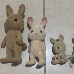Puede incluir: Colecci&oacute;n de cinco figuras de conejos de juguete de varios tama&ntilde;os. Los conejos tienen un pelaje suave y afelpado en tonos beige y marr&oacute;n, con orejas forradas de rosa y ojos negros. Un conejo lleva un atuendo azul y blanco.