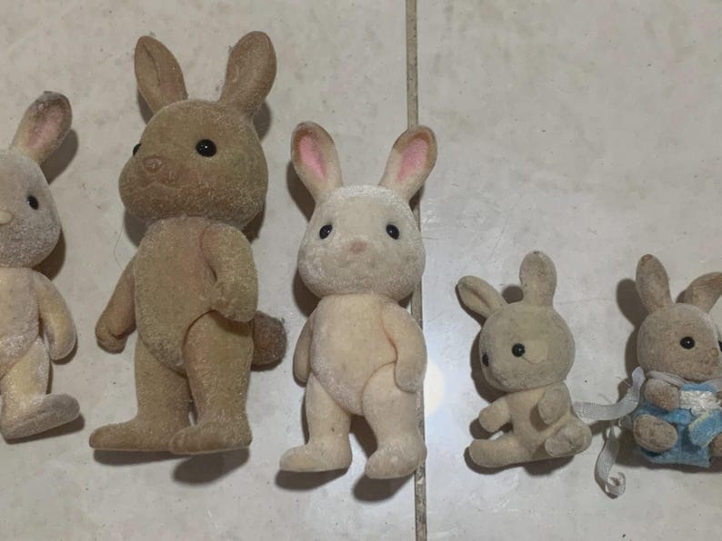 Puede incluir: Colecci&oacute;n de cinco figuras de conejos de juguete de varios tama&ntilde;os. Los conejos tienen un pelaje suave y afelpado en tonos beige y marr&oacute;n, con orejas forradas de rosa y ojos negros. Un conejo lleva un atuendo azul y blanco.