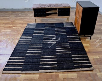 May include: A rectangular, dark blue rug with a geometric pattern of horizontal, off-white stripes. The rug is placed on a wooden floor, with a bench and cabinet in the background. The rug is labeled "Anaya Rugs Handmade Rugs".