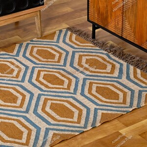 May include: Geometric patterned rug with a tan, blue, and off-white design. The rug features a repeating pattern of octagons and has a fringed edge. The rug is placed on a wooden floor.