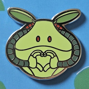 Könnte beinhalten: Ein grüner und silberner Emaille-Pin mit einer Cartoonfigur, die eine herzförmige Geste macht. Der Pin hat einen hellgrünen Körper, dunkelgrüne Akzente und rote Details. Der Pin ist vor einem blauen Hintergrund platziert.