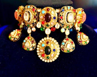 May include: A gold-toned choker necklace featuring large oval and circular medallions set with multicolored cabochon stones in red, blue, green, and white. The piece includes dangling teardrop-shaped pendants adorned with matching small colorful stones.