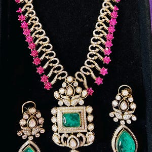 May include: A gold-toned jewelry set featuring a necklace and earrings. The necklace has a central emerald-cut green stone pendant, surrounded by diamonds. The earrings have teardrop-shaped green stones. Pink floral accents adorn the necklace.