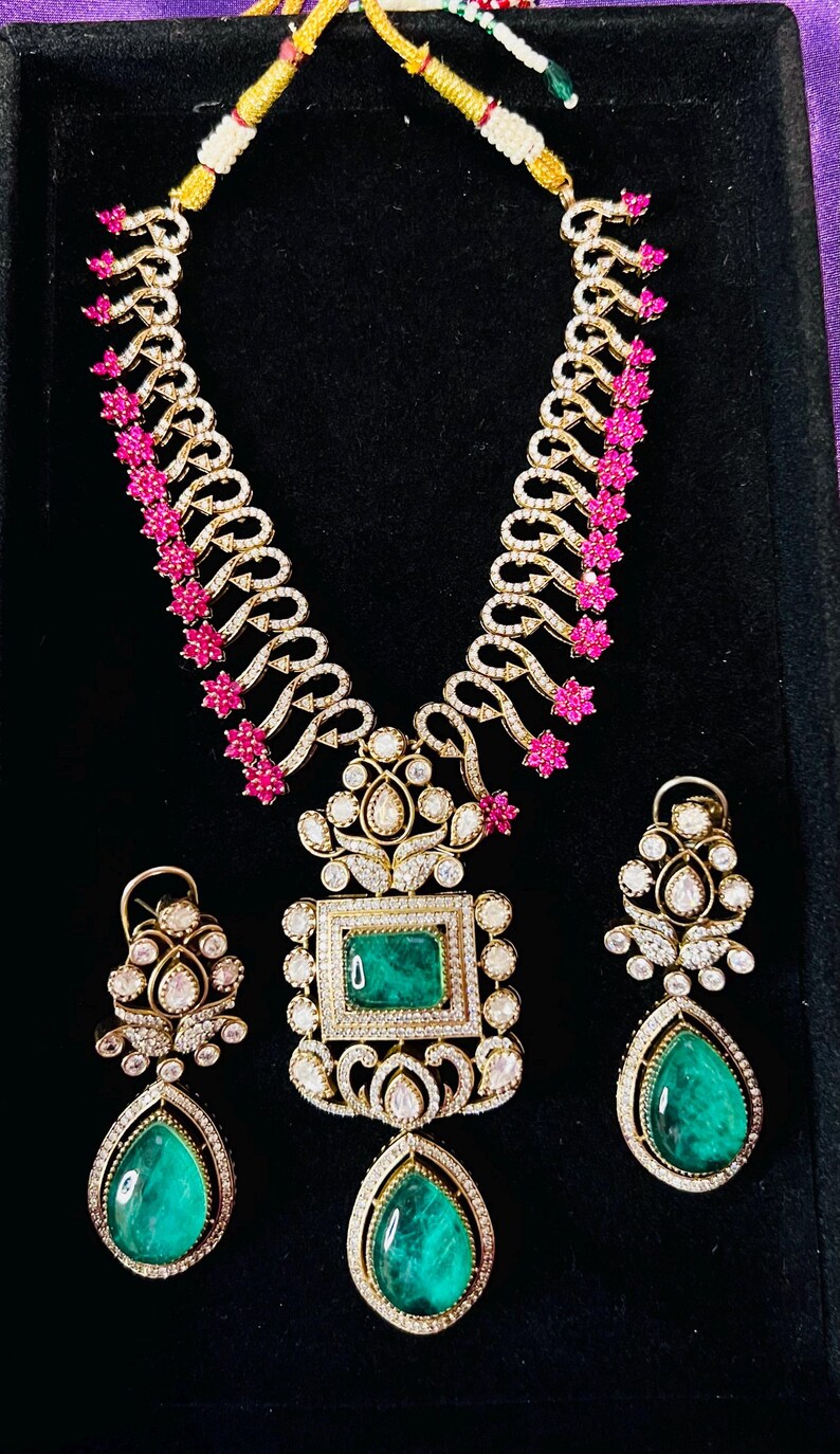 May include: A gold-toned jewelry set featuring a necklace and earrings. The necklace has a central emerald-cut green stone pendant, surrounded by diamonds. The earrings have teardrop-shaped green stones. Pink floral accents adorn the necklace.