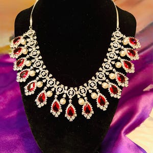 May include: A silver-toned necklace with a symmetrical design. The necklace features teardrop-shaped red gemstones, interspersed with small white pearls and clear crystals. The necklace is displayed on a black mannequin head against a purple background.