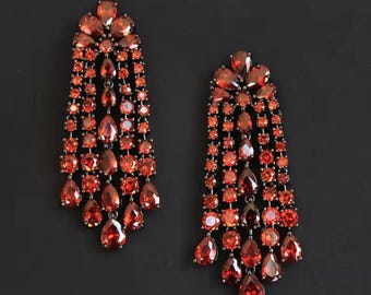 May include: A pair of elegant, dangling earrings. Each earring features a cascade of red-orange gemstones in various shapes, including teardrops and ovals, set on a dark metal frame. The earrings have a floral design at the top and a layered, fringed effect.