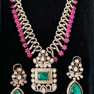 May include: A gold-toned necklace and earring set with emerald and diamond accents. The necklace features a large emerald pendant and is adorned with pink floral accents. The earrings have teardrop-shaped emeralds. The jewelry is set against a black background.
