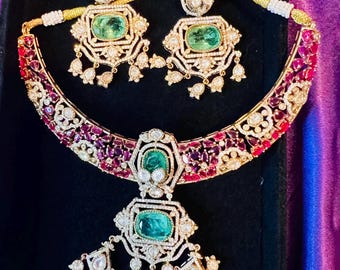 May include: A luxurious jewelry set featuring a necklace and matching earrings. The necklace and earrings have emerald-cut green gemstones, surrounded by smaller white stones and red accents. The set is displayed on a black surface.
