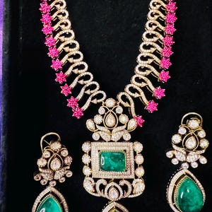May include: A gold-toned necklace and earring set featuring emerald green gemstones and sparkling clear stones. The necklace has a central square emerald pendant and a teardrop-shaped emerald pendant. The earrings have teardrop-shaped emeralds.