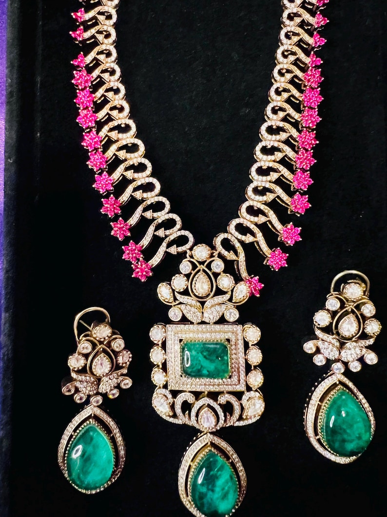 May include: A gold-toned necklace and earring set featuring emerald green gemstones and sparkling clear stones. The necklace has a central square emerald pendant and a teardrop-shaped emerald pendant. The earrings have teardrop-shaped emeralds.