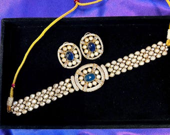 May include: A gold-toned jewelry set featuring a necklace and matching earrings. The necklace and earrings have a central blue stone surrounded by smaller white stones. The set is displayed on a black surface against a purple background.