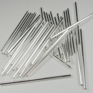 Op de afbeelding: Een stapel zilverkleurige metalen staven, ongeveer 100 in totaal, zijn willekeurig gerangschikt op een wit oppervlak. De staven zijn allemaal even lang en dik.