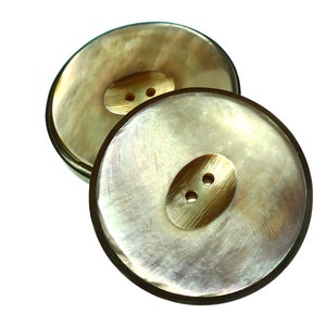 Può includere: Due grandi bottoni rotondi in madreperla iridescente con un design a due fori. I bottoni sono di colore beige chiaro con un leggero luccichio.