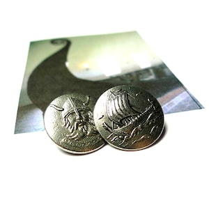 May include: Two silver buttons with Viking designs. One button shows a Viking face, and the other depicts a Viking ship sailing on the sea. A blurred image of a Viking ship is in the background.