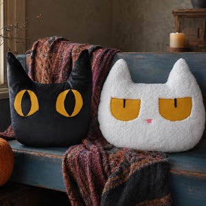 May include: Two cat-shaped throw pillows, one black with smooth fabric and one white with textured fleece, both featuring large yellow eyes with black pupils. The black pillow has pointed ears, while the white pillow has a small pink nose and pointed ears.