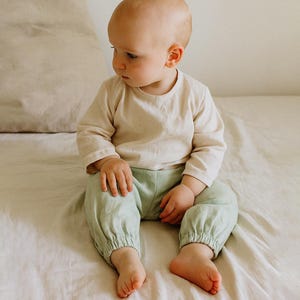May include: A baby wearing a long-sleeved beige shirt and light green pants with elastic cuffs sits on a bed. The baby is looking to the side. A beige pillow is in the background.