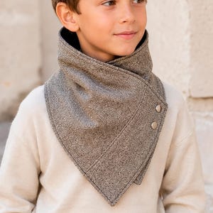 May include: A brown herringbone patterned neck warmer featuring a pointed front design and two metallic snap buttons on the side. The fabric has a textured weave, and the garment is worn around the neck over a cream-colored long-sleeve sweater.