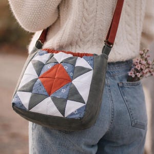 May include: A quilted crossbody bag with a geometric starburst pattern in shades of blue, white, gray, and orange. The bag has a dark green border and a brown strap. The bag is approximately 8 inches wide.