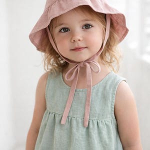 Può includere: Una bambina indossa un cappello da sole rosa chiaro con un nastro abbinato e un vestito verde chiaro senza maniche. Il cappello ha un'ampia tesa e il vestito ha un design semplice e arricciato.