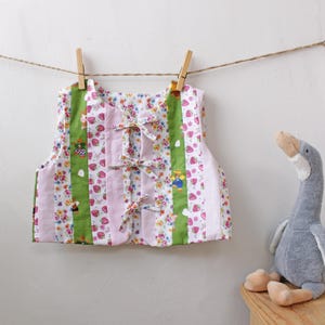 May include: A patterned vest with a floral and strawberry design, green stripes, and pink panels, hanging on a washing line with wooden pegs. A soft toy goose is positioned to the side. The vest is made of fabric.