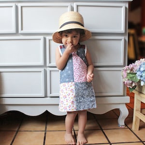 Puede incluir: Una niña pequeña con un sombrero de paja y un vestido de retazos se para frente a una cómoda blanca con cajones. El vestido está hecho de diferentes telas en tonos de rosa, azul y blanco.