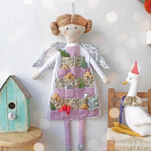 Puede incluir: Una muñeca de ángel de tela blanca con alas de purpurina plateada y un vestido a rayas rojas y blancas. El vestido tiene bolsillos que están llenos de pequeños objetos redondos marrones. La muñeca está de pie sobre un fondo blanco.