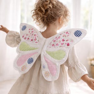 May include: A child wearing white butterfly wings with colourful patchwork details. The wings feature sections of pink stripes, green gingham, and strawberry print. Blue and floral patterned circles accent the wings. The child is wearing a beige dress.