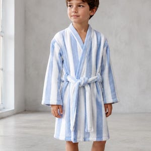 May include: A knee-length bathrobe featuring a vertical striped pattern in light blue and white. The robe has long sleeves, a shawl collar, and a matching fabric tie belt secured at the waist. The garment is made of a soft, textured fabric.