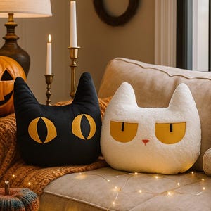 Peut inclure: Deux coussins en forme de chat, l'un noir avec des yeux dorés et l'autre blanc avec des yeux jaunes, sont posés sur un canapé beige. Une citrouille sculptée, des bougies et des citrouilles décoratives complètent le thème d'Halloween.