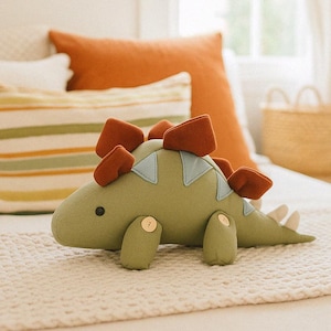 Puede incluir: Un peluche de dinosaurio verde con detalles marrones y azules. El juguete de peluche tiene placas marrones en la espalda, articulaciones de botón y un pequeño ojo negro. Descansa sobre una superficie texturizada crema, con almohadas en el fondo.