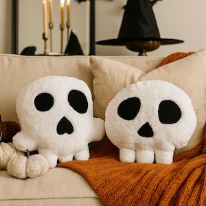 May include: Two white plush skull-shaped throw pillows with black felt eyes and nose details. The pillows feature a soft, textured fabric surface and are positioned alongside three small decorative pumpkins in brown, cream, and light beige colours.