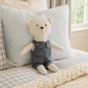 May include: A stuffed bear doll with a cream-colored body and a dark gray jumpsuit with white cross details. The bear is sitting on a bed with light blue and white bedding and pillows. The room has a neutral color palette.