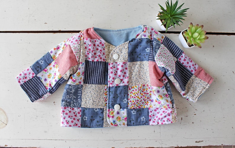 May include: A long-sleeved jacket featuring a patchwork design composed of various fabric squares in shades of blue, pink, white, and grey. The garment includes floral patterns, solid colors, and striped prints, finished with three white circular snap buttons.