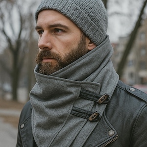 May include: A gray knit beanie and matching scarf with buckle closures. The scarf is layered and wraps around the neck. The person is wearing a black leather jacket.