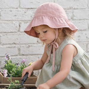 Op de afbeelding: Een kind met een lichtroze zonnehoed en een lichtgroene jurk is aan het tuinieren. Het kind gebruikt een kleine troffel en een handhark om een potplant te verzorgen. De achtergrond toont een witte bakstenen muur en een kleine plantenbak.