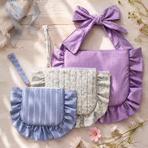 May include: Three fabric pouches with ruffled edges and wrist straps. The smallest is blue with white stripes, the middle is quilted with a blue floral pattern, and the largest is purple with a textured weave and a large tied bow at the top.