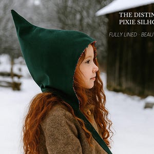 May include: A dark green pointed hood with a patterned lining. The text on the image reads: THE DISTINCTIVE PIXIE SILHOUETTE, FULLY LINED, BEAUTIFULLY FINISHED. The hood features a tall, structured peak and covers the head, extending down the neck.