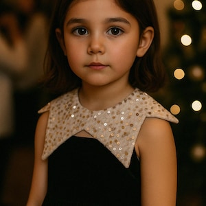 Könnte beinhalten: Ein junges Mädchen trägt ein schwarzes Kleid mit einem dekorativen Kragen. Der Kragen ist beige und mit goldenen Pailletten besetzt. Das Mädchen hat braune Haare und schaut in die Kamera. Im Hintergrund ist ein verschwommener Weihnachtsbaum zu sehen.