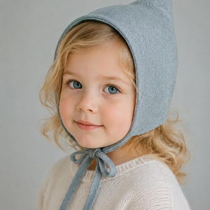 May include: A light blue pixie hat with a pointed top and chin ties. The text reads: PIXIE HAT PDF SEWING PATTERN 11 SIZES NEWBORN TO ADULT XL. The hat features a textured fabric construction that covers the head and secures with a bow under the chin.