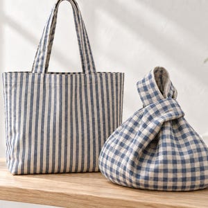 Puede incluir: Un bolso tote de rayas azules y blancas con dos asas junto a un bolso de nudo de cuadros vichy azules y blancos. Ambos bolsos están hechos de tela tejida y presentan un patrón clásico. El bolso tote es rectangular y el de nudo es redondeado.