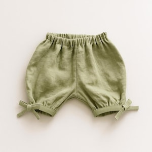 Puede incluir: Pantalones cortos de color verde salvia con cintura elástica y aberturas de pierna fruncidas. Cada pernera está rematada con un pequeño lazo de tela decorativo en el lado exterior, creando una silueta suave y redondeada para esta prenda lisa.