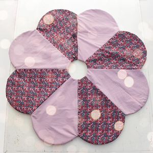 May include: A flower-shaped quilted trivet with alternating petals. Some petals feature a red floral pattern on a dark background, while others have a pink and white striped design. The trivet is designed to protect surfaces from heat.