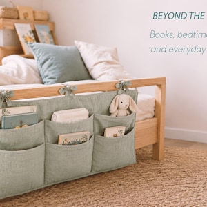 May include: A sage green fabric bedside organizer with three pockets, tied to a wooden bed frame with fabric ribbons. The pockets contain books and a small plush rabbit toy. Text on the wall reads: BEYOND THE NURSERY Books, bedtime favorites and everyday essentials.