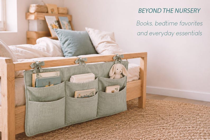 May include: A sage green fabric bedside organizer with three pockets, tied to a wooden bed frame with fabric ribbons. The pockets contain books and a small plush rabbit toy. Text on the wall reads: BEYOND THE NURSERY Books, bedtime favorites and everyday essentials.