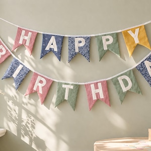 May include: A two-tiered fabric bunting banner spelling out HAPPY BIRTHDAY with white letters on swallowtail pennants. The flags feature a mix of solid pink, green, and yellow colors alongside blue and white floral patterns, all strung on white lace trim.