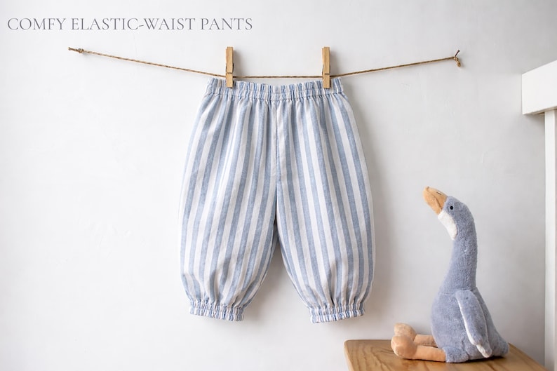May include: Pants with a light blue and white vertical striped pattern, featuring an elastic waistband and elasticized cuffs at the ankles. The text COMFY ELASTIC-WAIST PANTS is visible at the top left. A plush goose toy is positioned nearby.